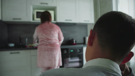 boy watches silently from foreground as woman in pink velvet robe prepares food at kitchen counter with oven, microwave, and red pot, illustrating quiet domestic moment filled with morning routine and stillness