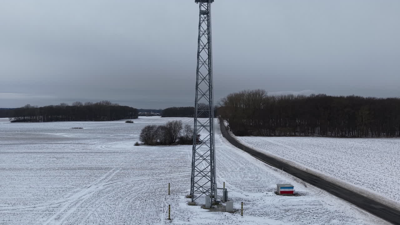 Aerial view of a modern mobile communication tower standing in a vast snow-covered winter landscape. The tall cellular mast with multiple antennas rises above frozen fields and distant forests