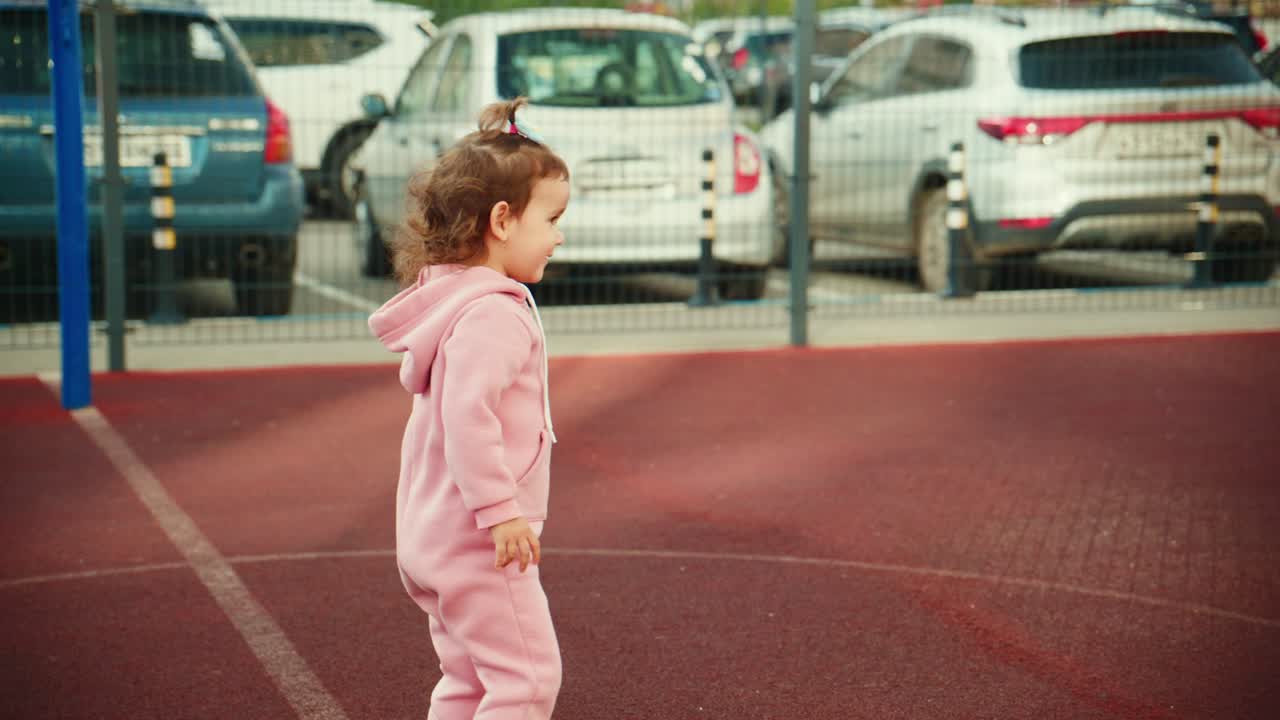 Un bambino piccolo in tuta rosa che gioca su una superficie colorata di un parco giochi, con delle auto sullo sfondo