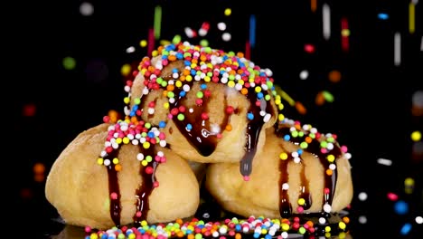 a stack of cream puffs with chocolate sauce is gradually covered in colorful rainbow sprinkles against a black background, under bright studio lighting, with a static camera