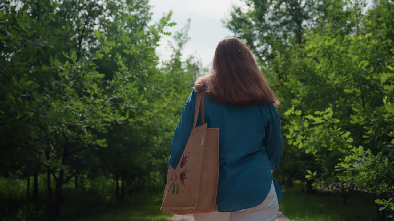 파란색 상의와 흰색 반바지를 입고 어깨에 자수 가방을 멘 채 녹색 나무들로 둘러싸인 공원 골목을 걷고 있는 통통한 학생의 뒷모습