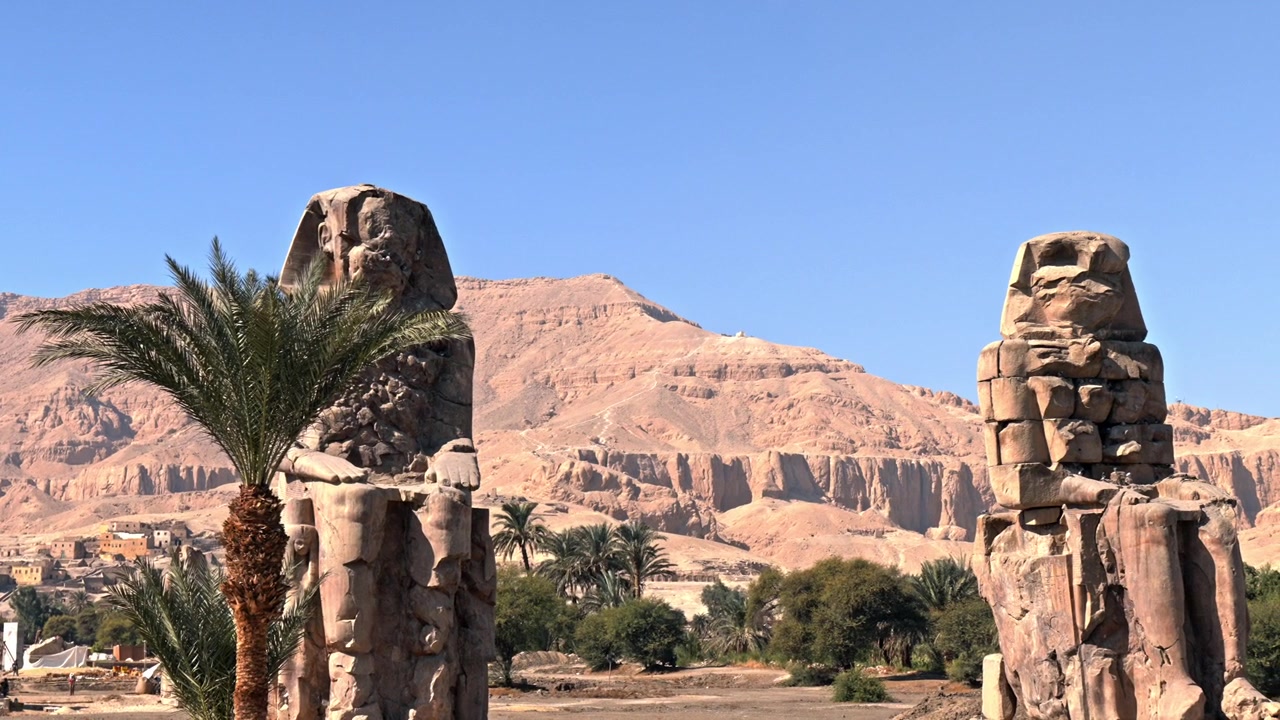 Sebuah bidikan lanskap lebar setinggi mata yang menangkap kedua patung monumental Colossi of Memnon di Tepi Barat Luxor, Mesir.
