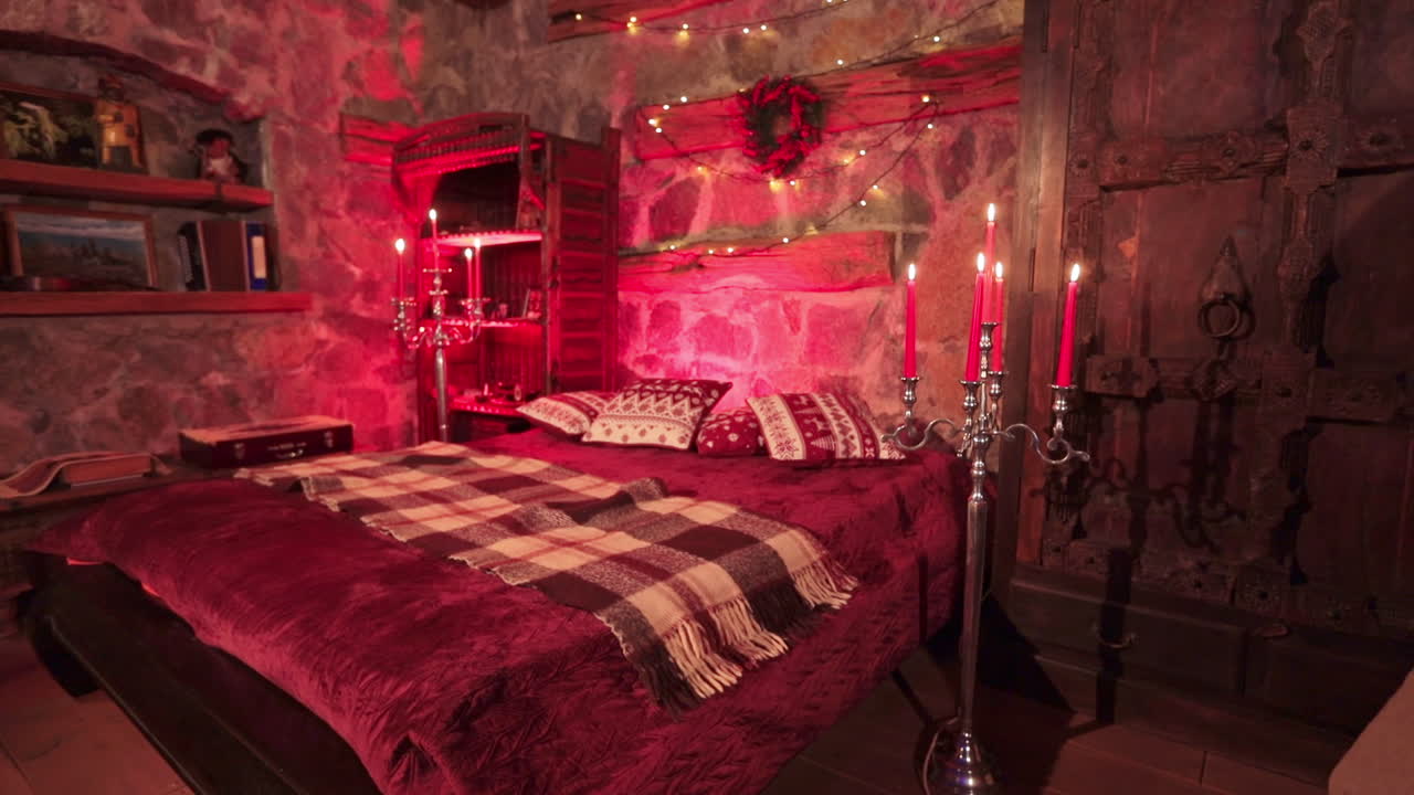 Big double bed in the beautifully decorated room with red lights. Stone walls, hard wooden old furniture and candles lighted in the countryside house bedroom.