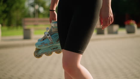 lady wearing black tights walking outdoors while holding cyan roller skates in her right hand, with blurred background featuring benches, lush greenery, and a flower pot