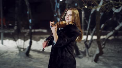 A talented girl in warm clothes is performing composition on the violin in a winter park in the evening on a blurred background. Camera motion to right