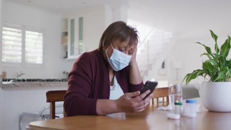 african american senior woman in face mask using smartphone at home, with medication on table