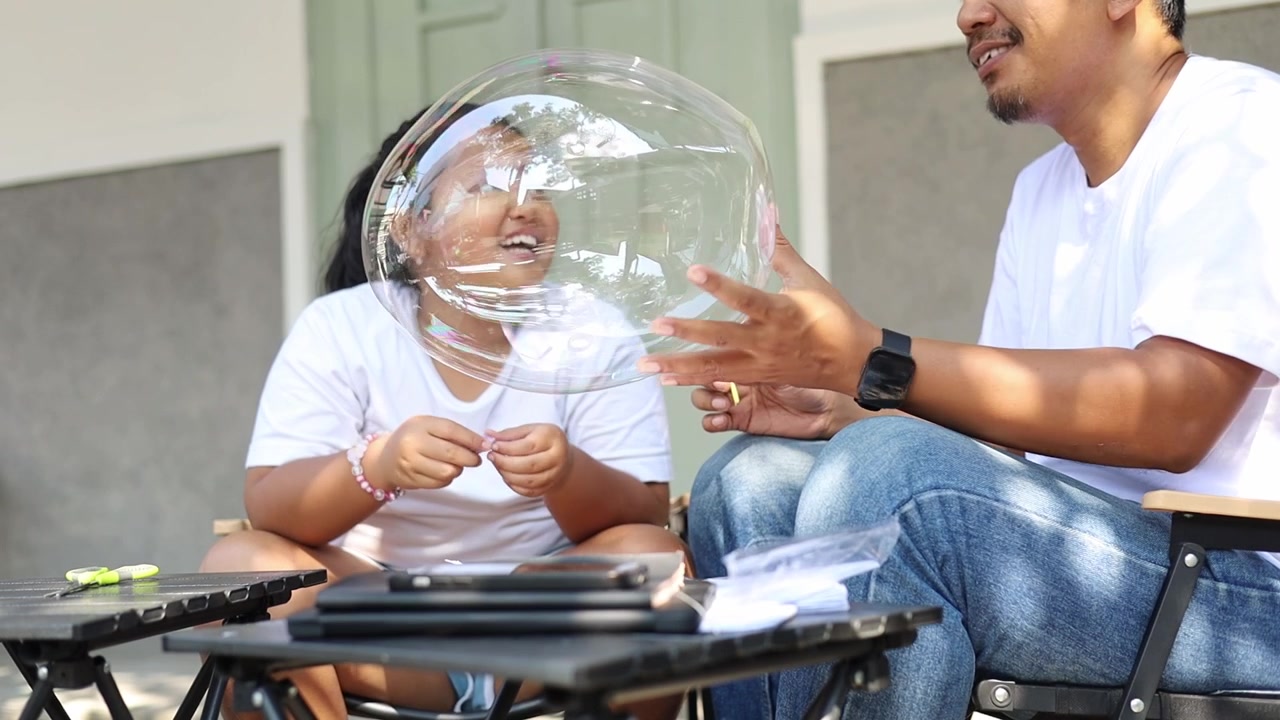 Father and daughter enjoying playful moments together by creating giant soap bubbles at home. Heartwarming family scene representing bonding, parenting, childhood happiness, learning through play