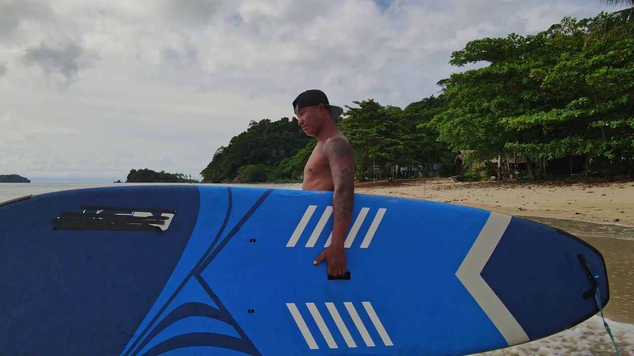 toma de seguimiento de un hombre asiático caminando con una tabla de remo a lo largo de una playa hacia el agua con playa y costa en tailandia