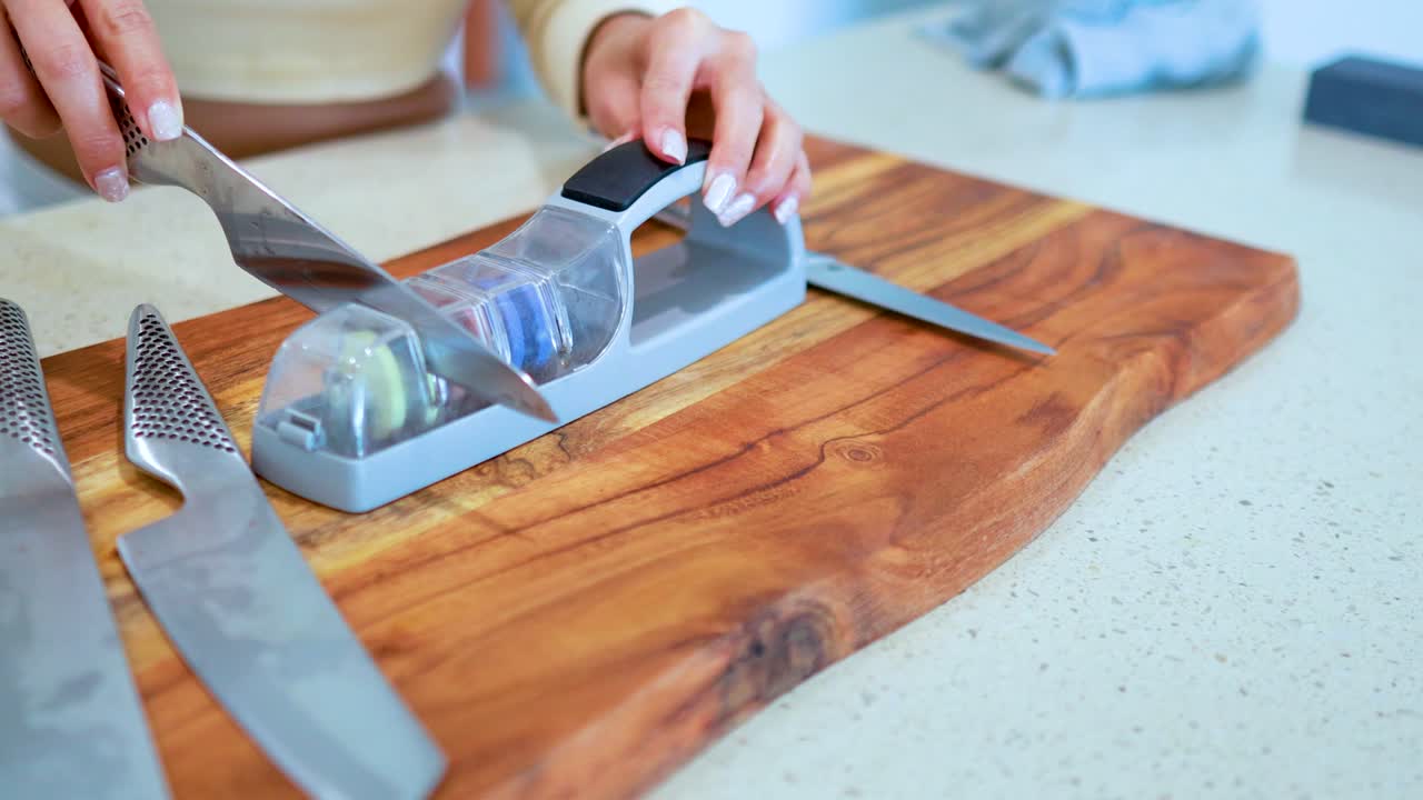 Hands sharpen a chef’s knife using a manual whetstone sharpener on a wooden cutting board in a brightly lit kitchen, with smooth camera movement