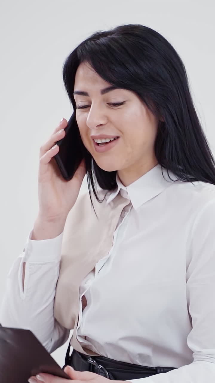 Attractive brunette talking the cellphone. Young woman in white blouse holding black folder and speaking on the mobile phone on the light studio background. Vertical video