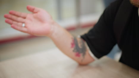 primer plano de la mano del hombre tatuado gesticulando en una conversación, con un fondo borroso que muestra el ambiente de la tienda minorista, la mano muestra un tatuaje y un anillo intrincados