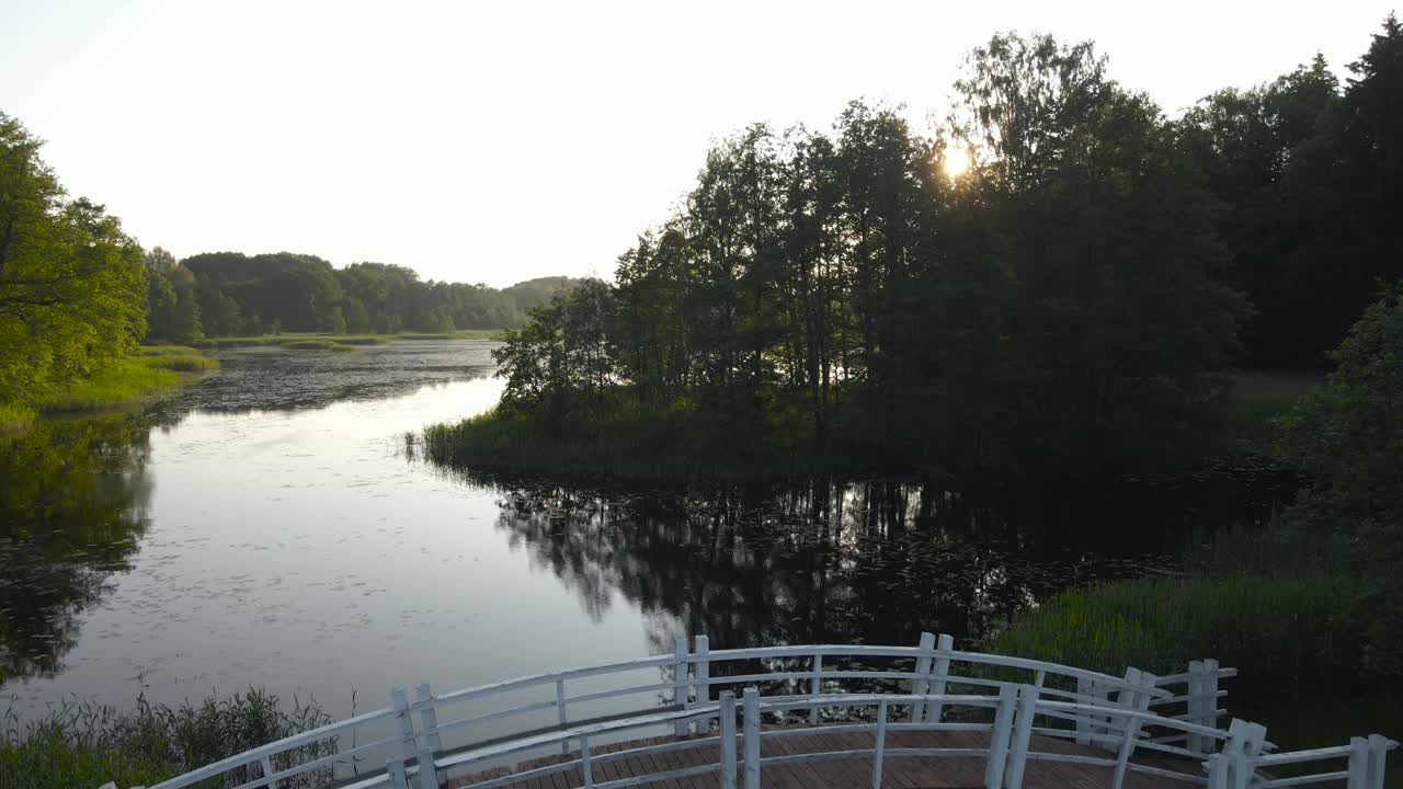 Aerial drone ascending view over wooden arch bridge across tranquil Alatskivi manor lake. Summer evening glow, setting sun through the trees. Scenic mirror water reflecting nature, romantic landscape