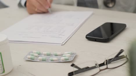 Arzt-Schreibt-Auf-Das-Weiße-Papier,-Zusammen-Mit-Seiner-Brille,-Seinem-Mobiltelefon-Und-Seiner-Medikamentenkapsel-Am-Tisch
