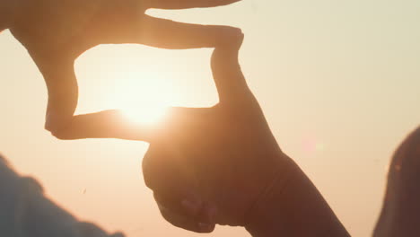rear view of lady framing glowing sun with fingertips in golden sunset sky, soft lens flare and hair lit by warm backlight, expressive hand gesture