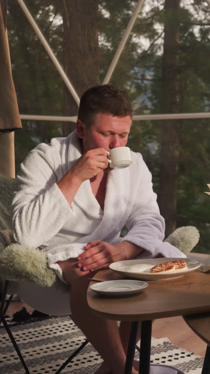 Guy drinks tea with woman cutting pancakes. Couple sits at table with vase with flowers near glass wall eating breakfast. Vacation in glamping house