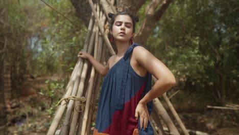 Eine-Frau-Mit-Dunklem-Haar-In-Einem-Blauen-Kleid-Steht-Neben-Einem-Baum-Und-Einem-Holzhaufen