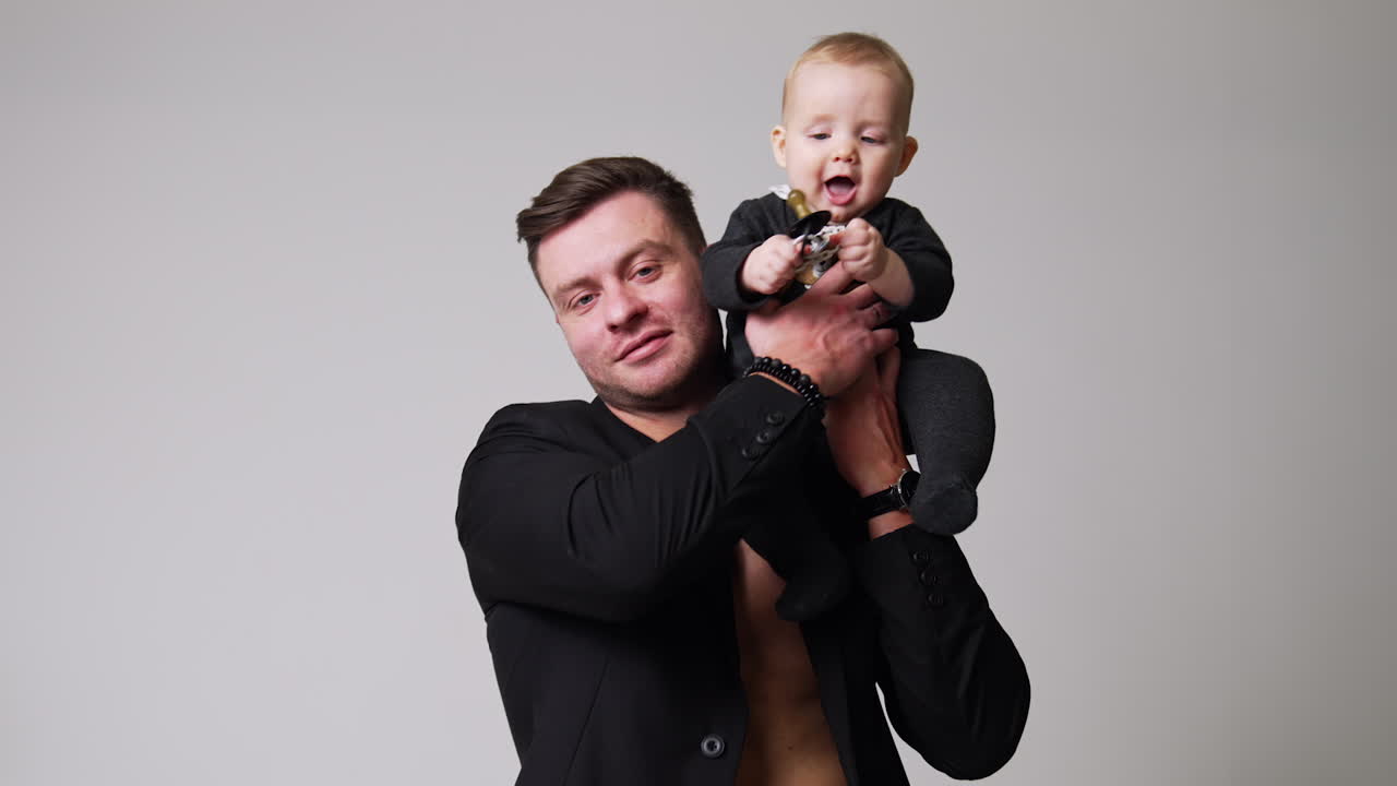 Handsome man in black jacket sits holding a baby. Father puts his lovely little child on the shoulder. White backdrop.