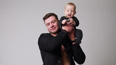 handsome man in black jacket sits holding a baby. father puts his lovely little child on the shoulder. white backdrop.