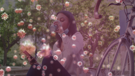 Multiple-covid-19-cells-floating-against-woman-with-lowered-face-mask-drinking-coffee-in-the-park