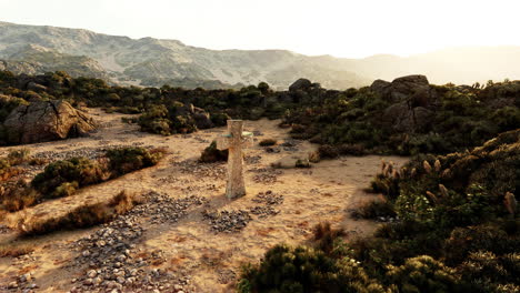 una cruz de piedra se alza en una ladera desértica con montañas en el fondo