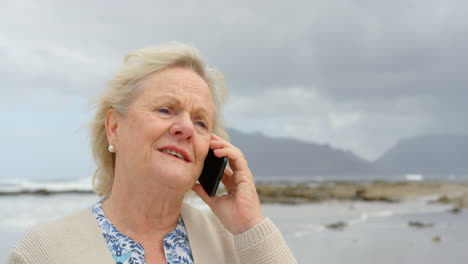 Front-view-of-old-caucasian-senior-woman-talking-on-mobile-phone-at-beach-4k-