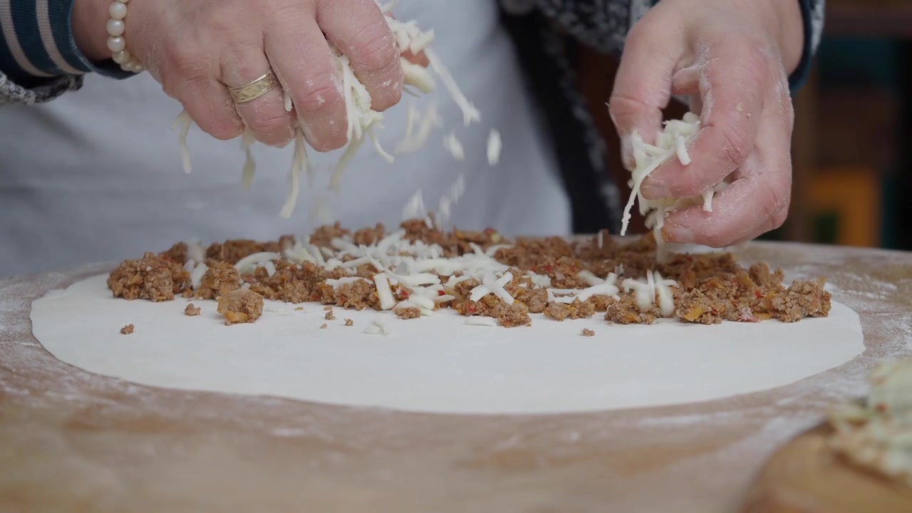 Primo piano di mani che spargono formaggio grattugiato su un impasto con ripieno di carne macinata