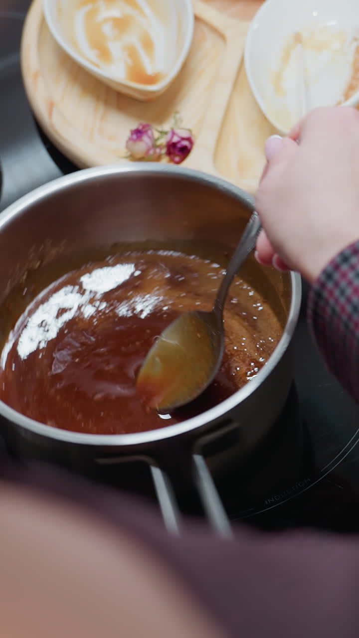 vista parcial de una persona agitando sopa gruesa en una olla de acero inoxidable en una estufa de inducción mientras ajusta su posición, con bandeja de madera, cuencos usados y utensilios de cocina dispuestos en mármol oscuro
