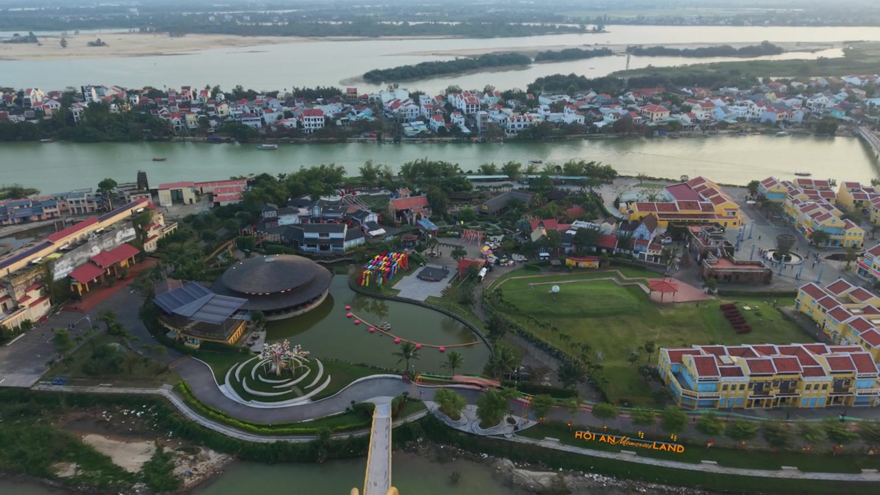 Màn hình dọc: Toàn cảnh từ trên cao khu nghỉ dưỡng Hội An ven sông lúc bình minh yên tĩnh, khu phức hợp nhà hát hình tròn, bãi cỏ được cắt tỉa gọn gàng, cầu đi bộ, mặt nước phản chiếu nhẹ nhàng, đường chân trời thị trấn xa xa, ánh vàng dịu
