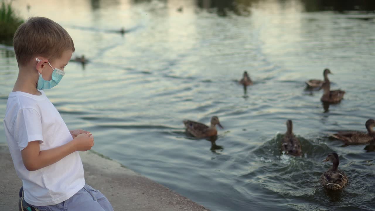 의료 마스크를 입은 소년이 호수에서 오리에게 먹이를 주고 있다.