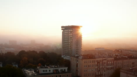 Slide-and-pan-shot-of-apartment-house-towering-above-other-town-development.-Tall-prefab-against-sun-rising-behind-fog.-Warsaw,-Poland