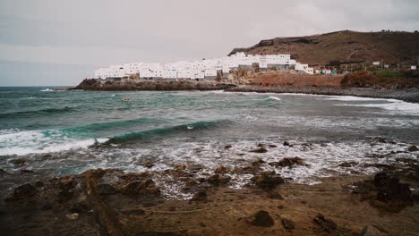 Slow-motion-pan-of-crashing-waves-on-rocky-shore-with-beautiful-white-buildings-on-hill-in-background