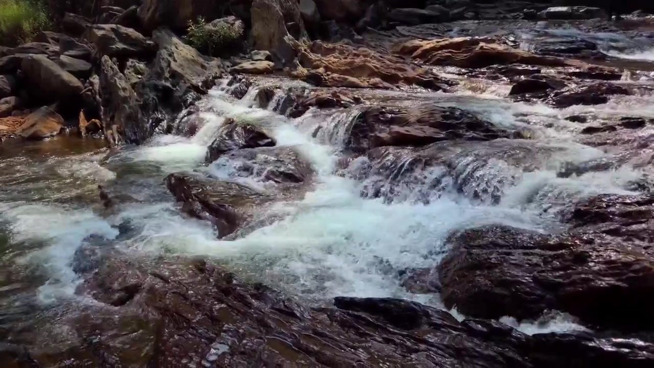 Un primo piano dinamico di acqua cristallina che scorre impetuosa su pietre di fiume scure e bagnate. La luce del sole colpisce l'acqua bianca e turbolenta, creando una texture scintillante e ad alta energia, perfetta per la natura e il benessere.