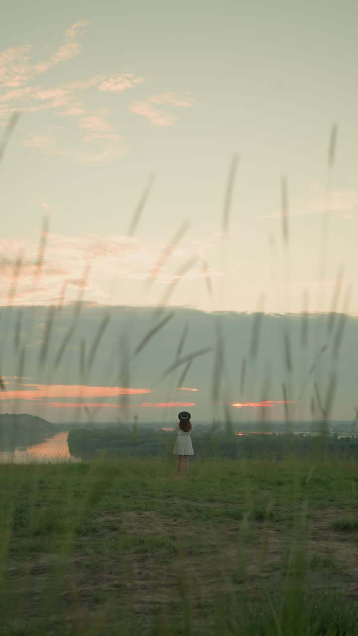 una mujer con un vestido blanco y un sombrero se encuentra sola en un campo de hierba aislado junto a un lago tranquilo al atardecer. ella mira el entorno sereno, abrazando el momento pacífico y reflexivo