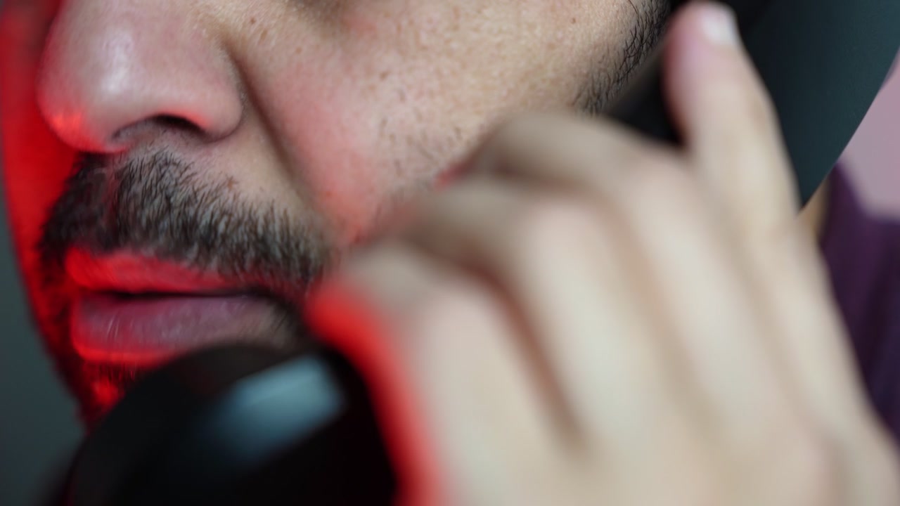 An extreme close-up shot of a man with a stubble beard holding a black landline telephone receiver to his face. The dramatic indoor scene is illuminated by intense red studio lighting.