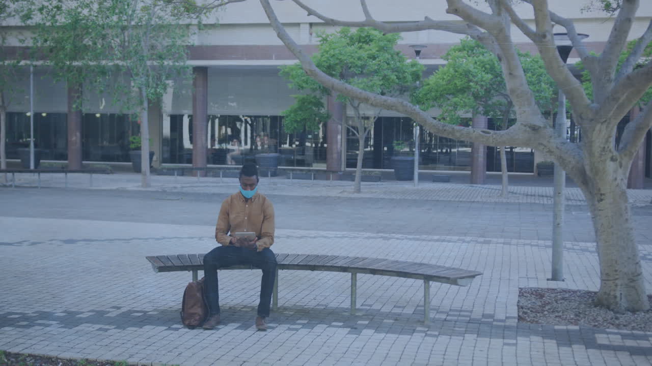 Using tablet under tree, man wearing mask sitting on bench outdoors