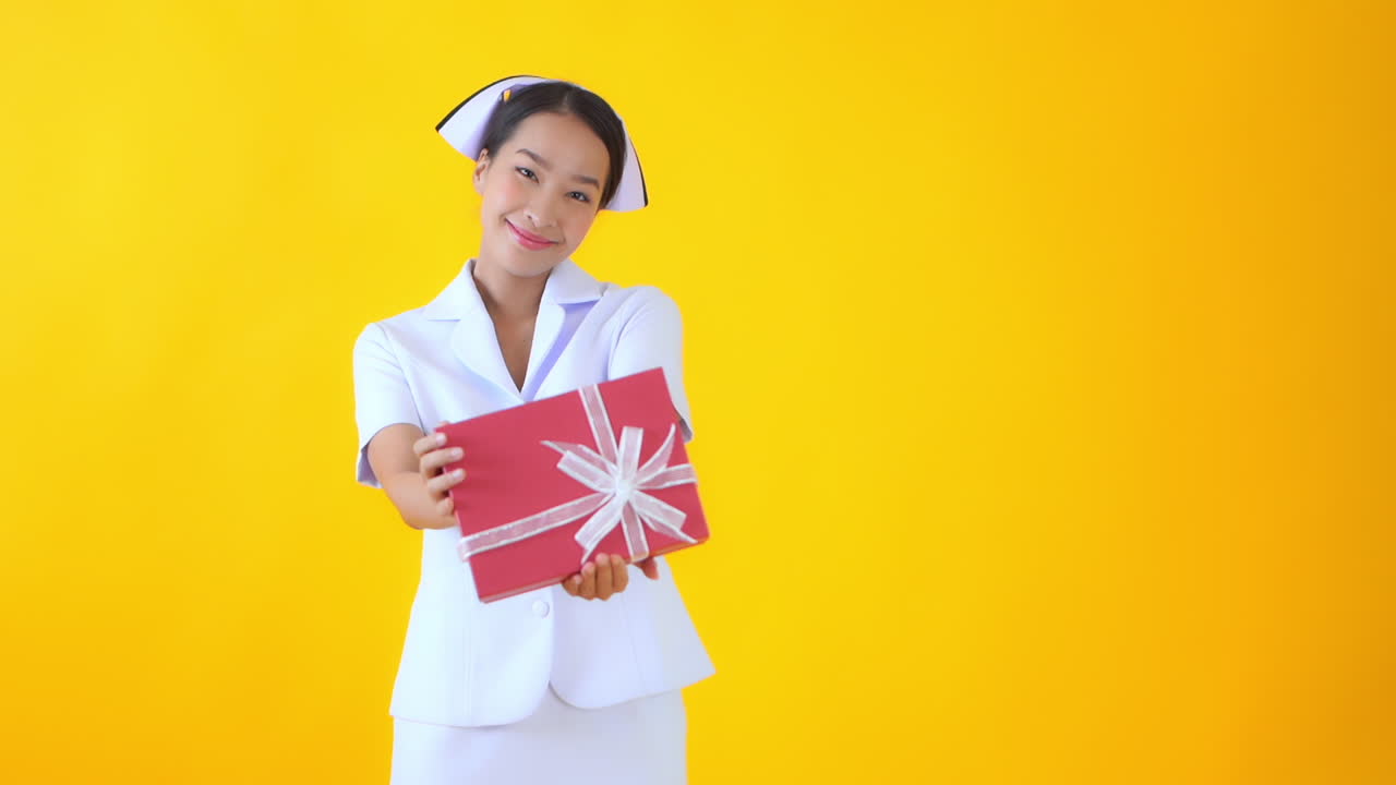 Static slow-motion shot of a cute Asian nurse holding a present in her hands and smiling cheerfully in front of a yellow background