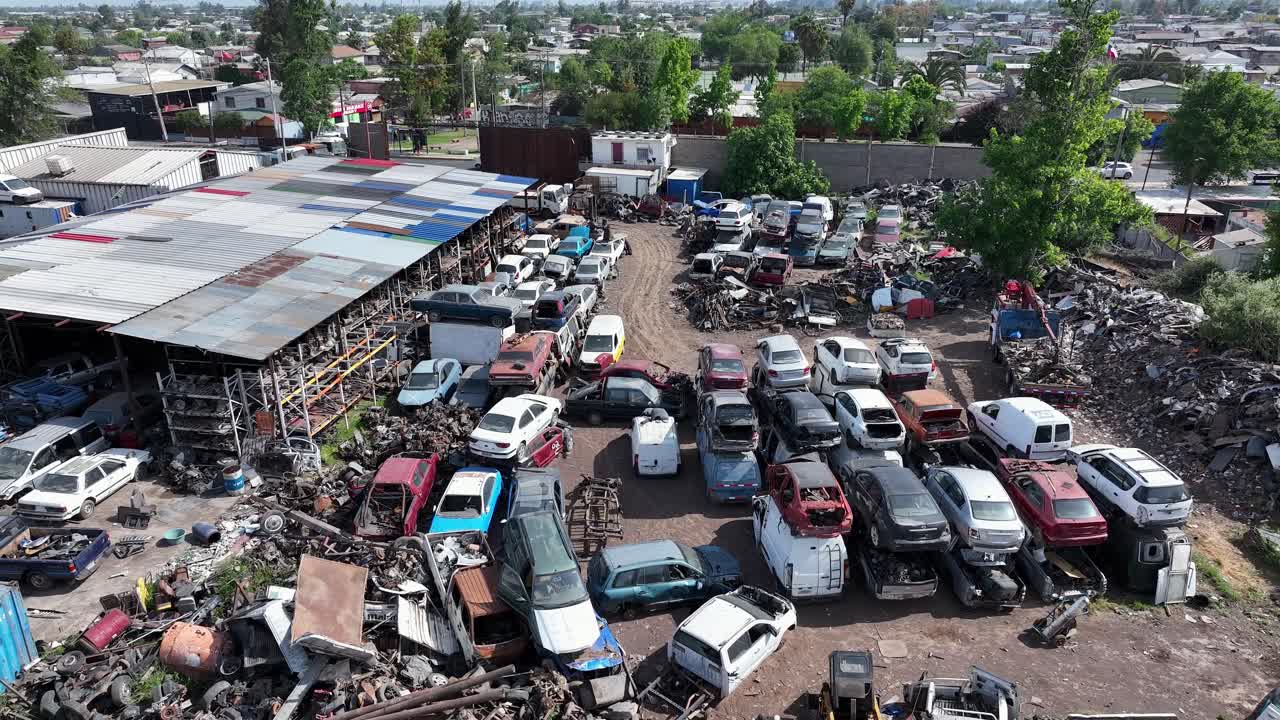 Forward drone aerial of junkyard with rows of scrapped cars, warehouse structure, and residential neighborhood with trees backdrop