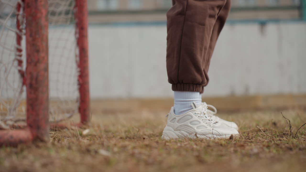 Primer plano de pies caucásicos cerca del poste de la portería sobre hierba, zapatillas blancas y calcetines que pisan con cautela junto a la red oxidada, puño de chándal marrón, suelo otoñal, ambiente de patio de recreo urbano, composición en ángulo bajo que transmite