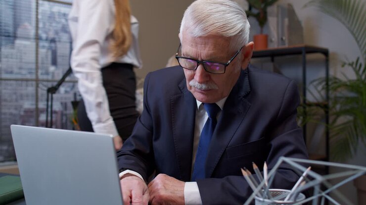 Focused senior business man entrepreneur typing on laptop doing research, sitting at office desk