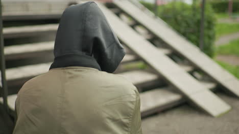 rear view of kid in hooded jacket holding handheld device while playing game, adjusting posture outdoors near staircase, focus on casual moment with muted urban background and red brick building