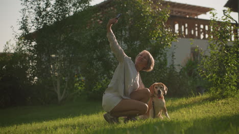 mujer tomando una selfie con su perro centrado en algo más al aire libre en un jardín verde exuberante bajo un clima soleado, con edificios y árboles en el fondo, el perro está sentado, jadeando, mientras el dueño sonríe