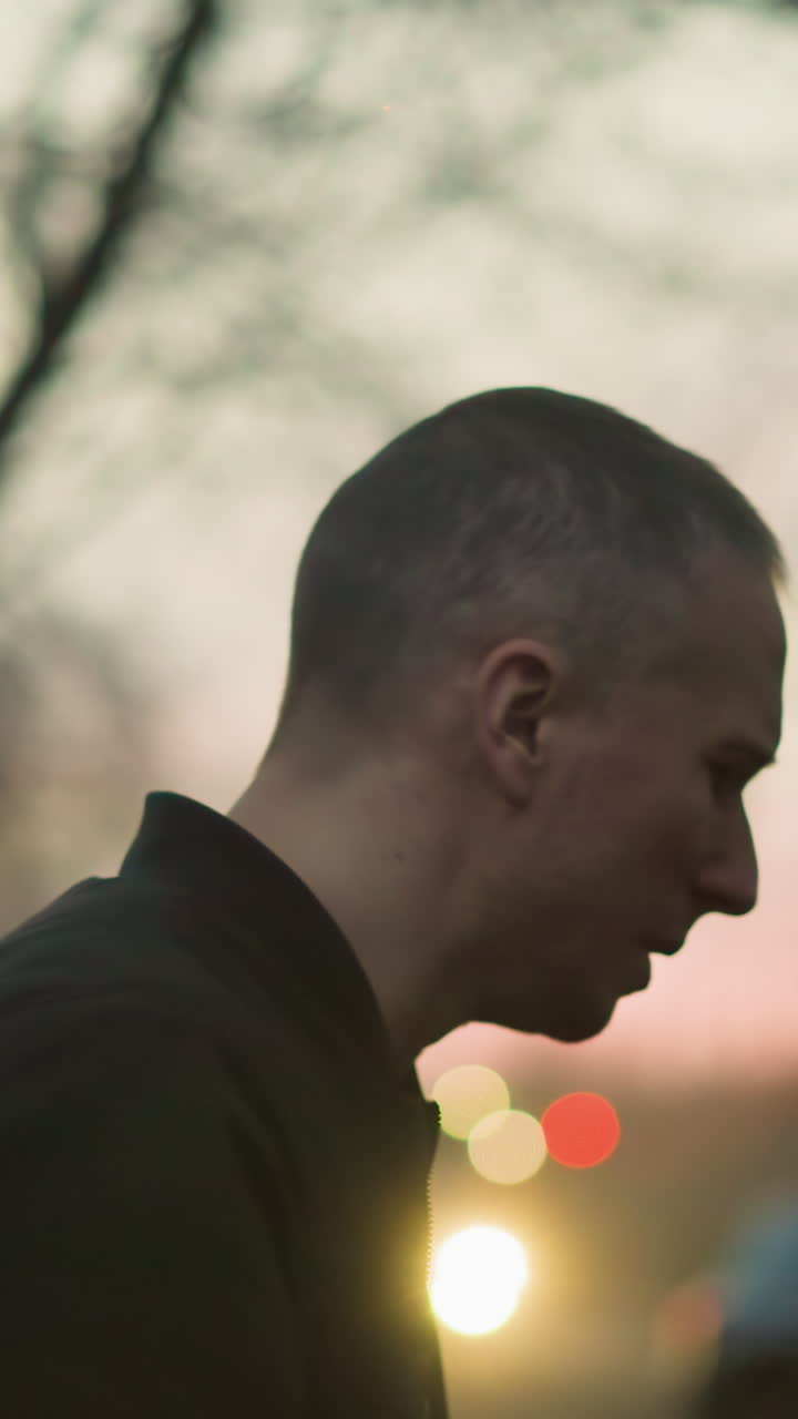 un hombre con una chaqueta verde fumando un cigarrillo al aire libre al anochecer, el fondo está borroso con luces suaves y cálidas