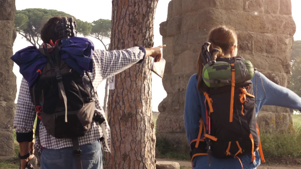una joven pareja de mochileros turistas caminando con una guía de mapas cerca del acueducto romano arcos en el parco degli acquedotti ruinas del parque en roma al amanecer con guitarra y saco de dormir cámara estacionaria de cámara lenta