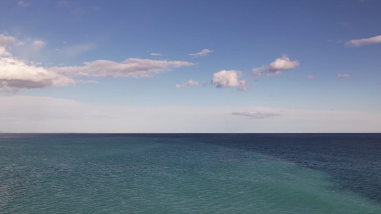 Vast expanse of turquoise and deep blue ocean stretches to the horizon under a bright sky dotted with soft white clouds.