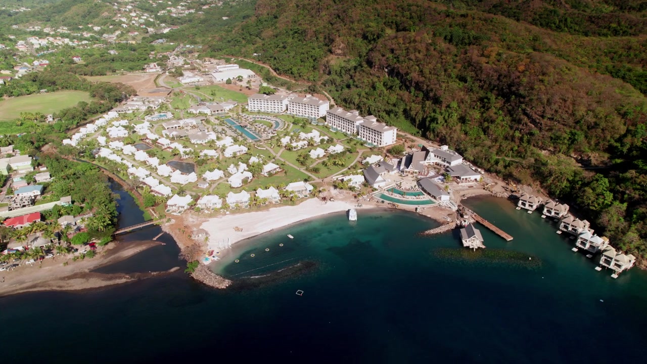 Lüks bir Karayip tatil köyünün villa kümelerini, merkezi havuz kompleksini, sahil şeridini ve çevresindeki tropikal dağ arazisini sergileyen yüksek irtifadan geniş açılı bir drone çekimi.
