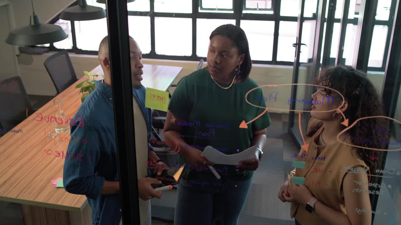 Diverse coworkers brainstorming at glass wall after man pointing at diagram, placing sticky notes