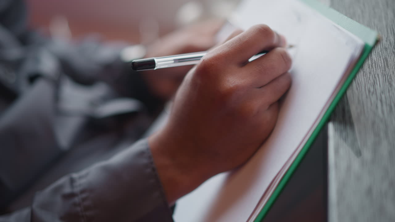 Immigrant writing with pen on white paper clipped to green folder near window, close-up of hands and focus on movement of pen, suggesting concentration, creativity, or preparation in calm indoor setting