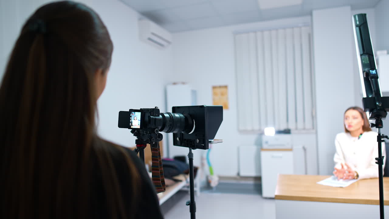 Rear view of a brunette girl standing in front of the camera recording video. Female blogger sitting at desk speaks to camera at blurred backdrop.