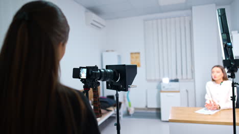rear view of a brunette girl standing in front of the camera recording video. female blogger sitting at desk speaks to camera at blurred backdrop.