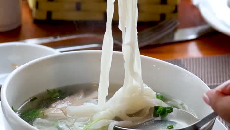 Close-up of noodles being lifted from a bowl with chopsticks and a spoon.